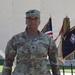 A U.S. Army Captain named Gillis stands at attention in Army Combat Uniform (ACU/OCP pattern) in front of a white architectural structure. The officer displays Captain rank insignia (two silver bars) on both the patrol cap and chest. Multiple military flags, including the U.S. flag and what appears to be a unit guidon with gold fringe, are visible in the background.