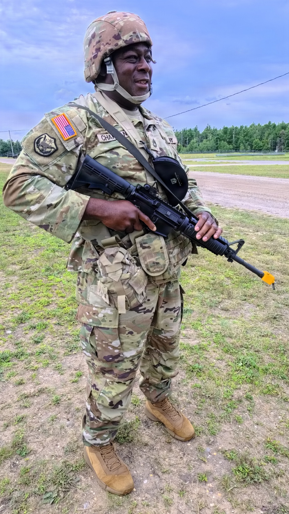 A U.S. Army soldier stands outdoors on a grassy training area wearing full Operational Camouflage Pattern (OCP) uniform and combat helmet. The soldier is holding an M16-style rifle with a yellow training muzzle cap attached, indicating a training exercise. Visible patches and name tape suggest an active-duty or National Guard affiliation.