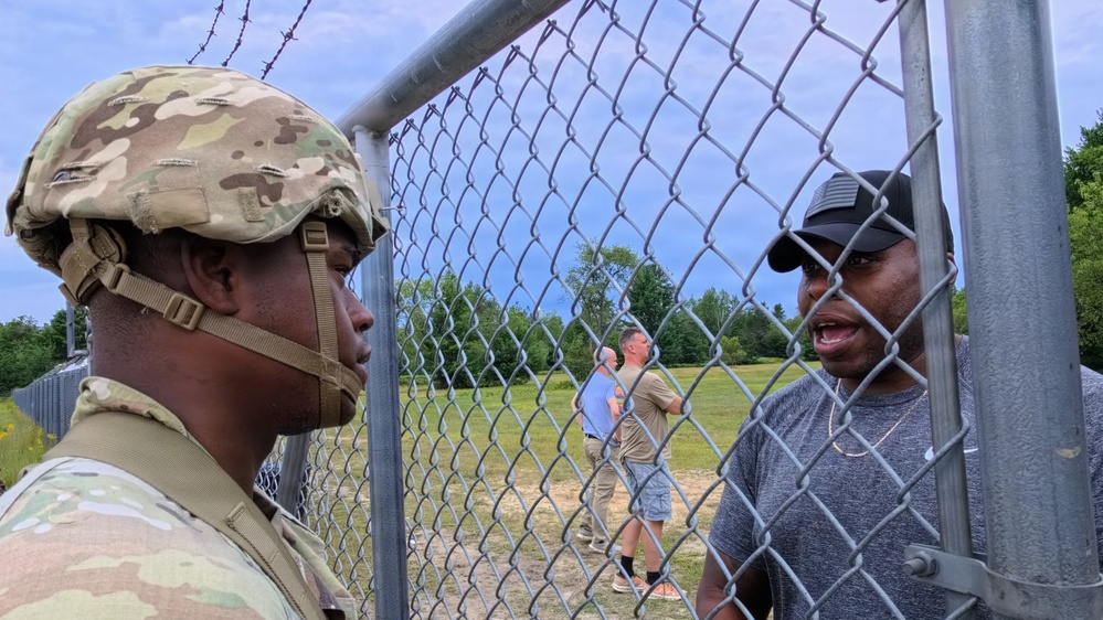 A soldier in full combat uniform and helmet stands on one side of a chain-link fence topped with barbed wire, facing a civilian man in a gray t-shirt and black cap on the other side. The two appear to be engaged in a conversation or interaction at what appears to be a secured perimeter gate. In the background, two other individuals can be seen in an open grassy area.