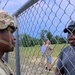 A soldier in full combat uniform and helmet stands on one side of a chain-link fence topped with barbed wire, facing a civilian man in a gray t-shirt and black cap on the other side. The two appear to be engaged in a conversation or interaction at what appears to be a secured perimeter gate. In the background, two other individuals can be seen in an open grassy area.