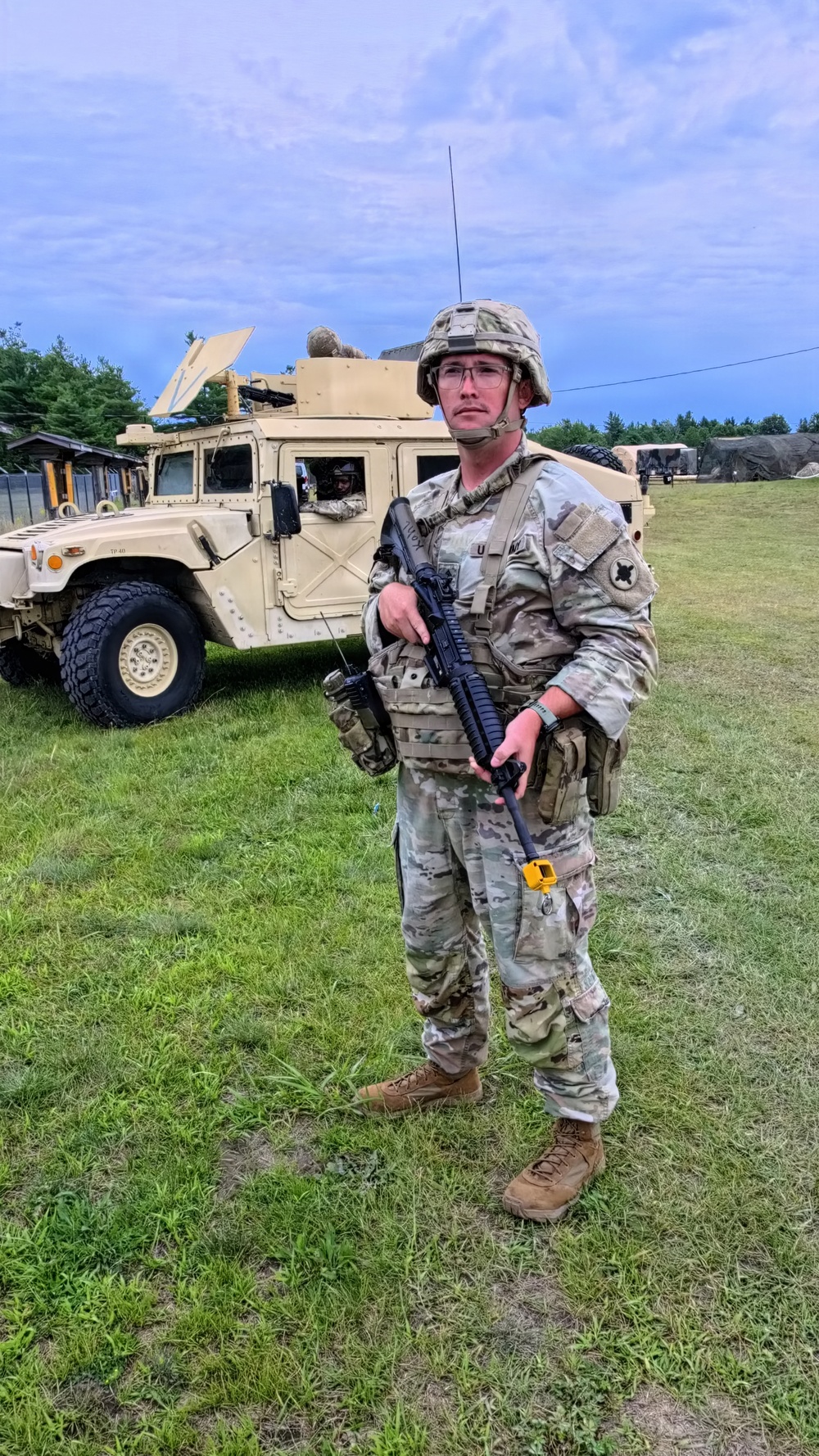 A U.S. Army soldier in full combat gear stands on a grassy field during what appears to be a military training exercise. The soldier is equipped with OCP (Operational Camouflage Pattern) uniform, plate carrier, ACH helmet, and is holding an M4-style carbine with a yellow training device attached to the muzzle. A tan HMMWV (Humvee) with a satellite dish and another soldier visible in the turret is positioned in the background.