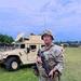 A U.S. Army soldier in full combat gear stands on a grassy field during what appears to be a military training exercise. The soldier is equipped with OCP (Operational Camouflage Pattern) uniform, plate carrier, ACH helmet, and is holding an M4-style carbine with a yellow training device attached to the muzzle. A tan HMMWV (Humvee) with a satellite dish and another soldier visible in the turret is positioned in the background.