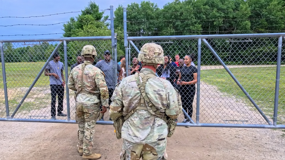 Two soldiers in U.S. Army OCP (Operational Camouflage Pattern) uniforms and combat helmets stand facing a chain-link gate where a group of civilians is gathered on the other side. The setting appears to be an outdoor area with a grassy field and trees in the background, surrounded by a chain-link fence topped with barbed wire. This appears to be a training exercise or controlled access scenario.