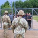 Two soldiers in U.S. Army OCP (Operational Camouflage Pattern) uniforms and combat helmets stand facing a chain-link gate where a group of civilians is gathered on the other side. The setting appears to be an outdoor area with a grassy field and trees in the background, surrounded by a chain-link fence topped with barbed wire. This appears to be a training exercise or controlled access scenario.