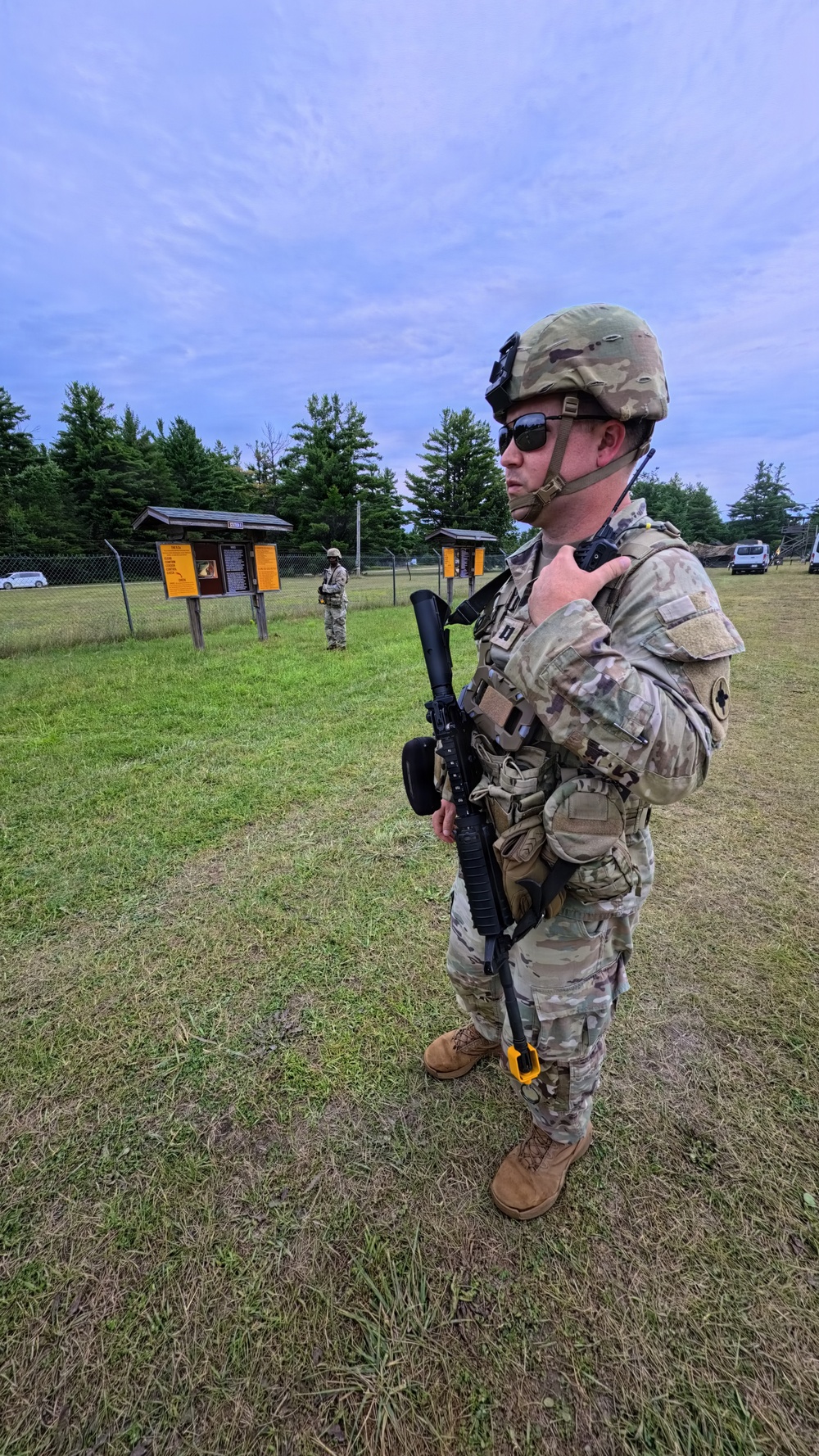 A U.S. Army soldier in full combat gear stands on a grassy field, holding a rifle and appearing to communicate via radio. A second soldier is visible in the background near informational bulletin boards. The setting appears to be a military training area or installation.