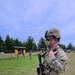 A U.S. Army soldier in full combat gear stands on a grassy field, holding a rifle and appearing to communicate via radio. A second soldier is visible in the background near informational bulletin boards. The setting appears to be a military training area or installation.