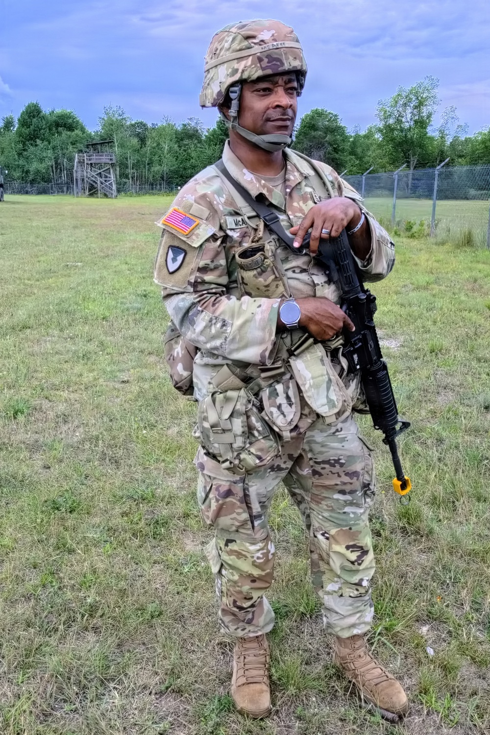 A U.S. Army soldier in full Operational Camouflage Pattern (OCP) uniform stands on a grassy field at what appears to be a military training area. The soldier is equipped with a combat helmet, body armor, tactical vest with pouches, and is holding an M4-style carbine rifle. A wooden observation tower and chain-link fence are visible in the background.
