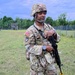 A U.S. Army soldier in full Operational Camouflage Pattern (OCP) uniform stands on a grassy field at what appears to be a military training area. The soldier is equipped with a combat helmet, body armor, tactical vest with pouches, and is holding an M4-style carbine rifle. A wooden observation tower and chain-link fence are visible in the background.