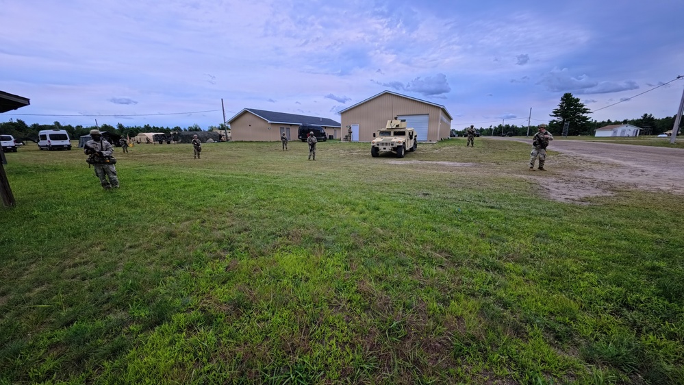 The image shows military personnel conducting what appears to be a training exercise in an open grassy area near several buildings. Multiple soldiers in camouflage uniforms are dispersed across the field, with at least one HMMWV (Humvee) military vehicle visible in the background. The setting appears to be a military training installation or mock village environment.