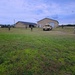 The image shows military personnel conducting what appears to be a training exercise in an open grassy area near several buildings. Multiple soldiers in camouflage uniforms are dispersed across the field, with at least one HMMWV (Humvee) military vehicle visible in the background. The setting appears to be a military training installation or mock village environment.