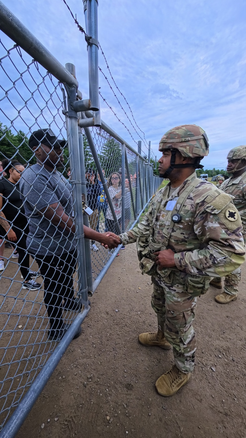 A U.S. Army soldier in full OCP (Operational Camouflage Pattern) uniform and combat helmet is shaking hands with a civilian through a chain-link fence topped with barbed wire. At least one additional soldier is visible in the background. The setting appears to be a military installation or training area during what may be a public engagement or family day event.