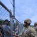 A U.S. Army soldier in full OCP (Operational Camouflage Pattern) uniform and combat helmet is shaking hands with a civilian through a chain-link fence topped with barbed wire. At least one additional soldier is visible in the background. The setting appears to be a military installation or training area during what may be a public engagement or family day event.