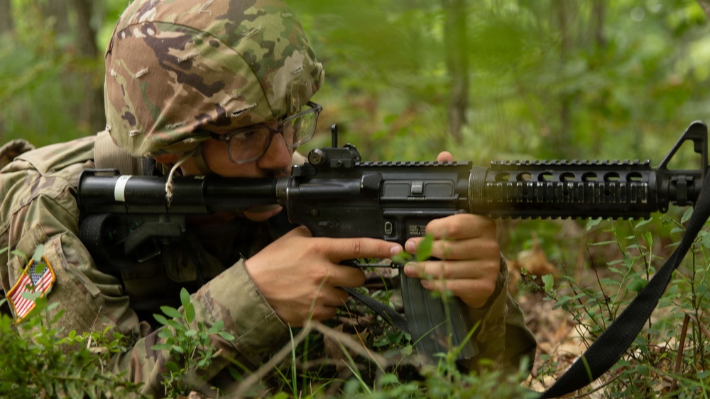A U.S. Army soldier in MultiCam/OCP camouflage uniform is in a prone or low position in a wooded area, aiming an M4 carbine with an attached M203 grenade launcher. The soldier is wearing glasses and a patrol cap under their helmet cover, with a U.S. flag patch visible on the left shoulder. The setting appears to be a field training exercise in a forested environment.
