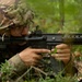 A U.S. Army soldier in MultiCam/OCP camouflage uniform is in a prone or low position in a wooded area, aiming an M4 carbine with an attached M203 grenade launcher. The soldier is wearing glasses and a patrol cap under their helmet cover, with a U.S. flag patch visible on the left shoulder. The setting appears to be a field training exercise in a forested environment.