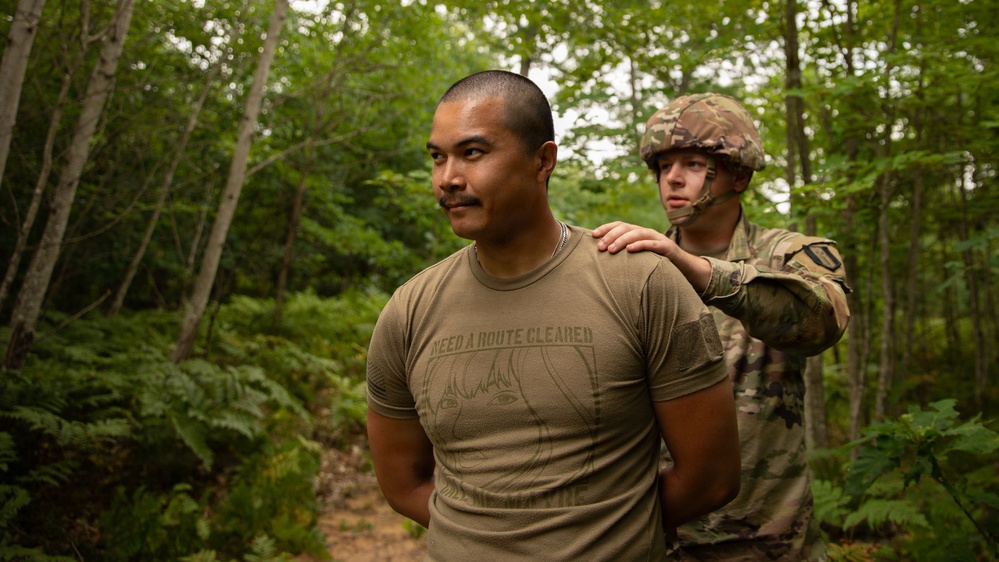 Two individuals are standing on a forest trail. One person in civilian attire wearing an olive/tan t-shirt with text and graphic print stands in the foreground, while a uniformed soldier in MultiCam/OCP camouflage with a combat helmet stands behind, placing a hand on the civilian's shoulder. The setting appears to be a wooded training or exercise environment.