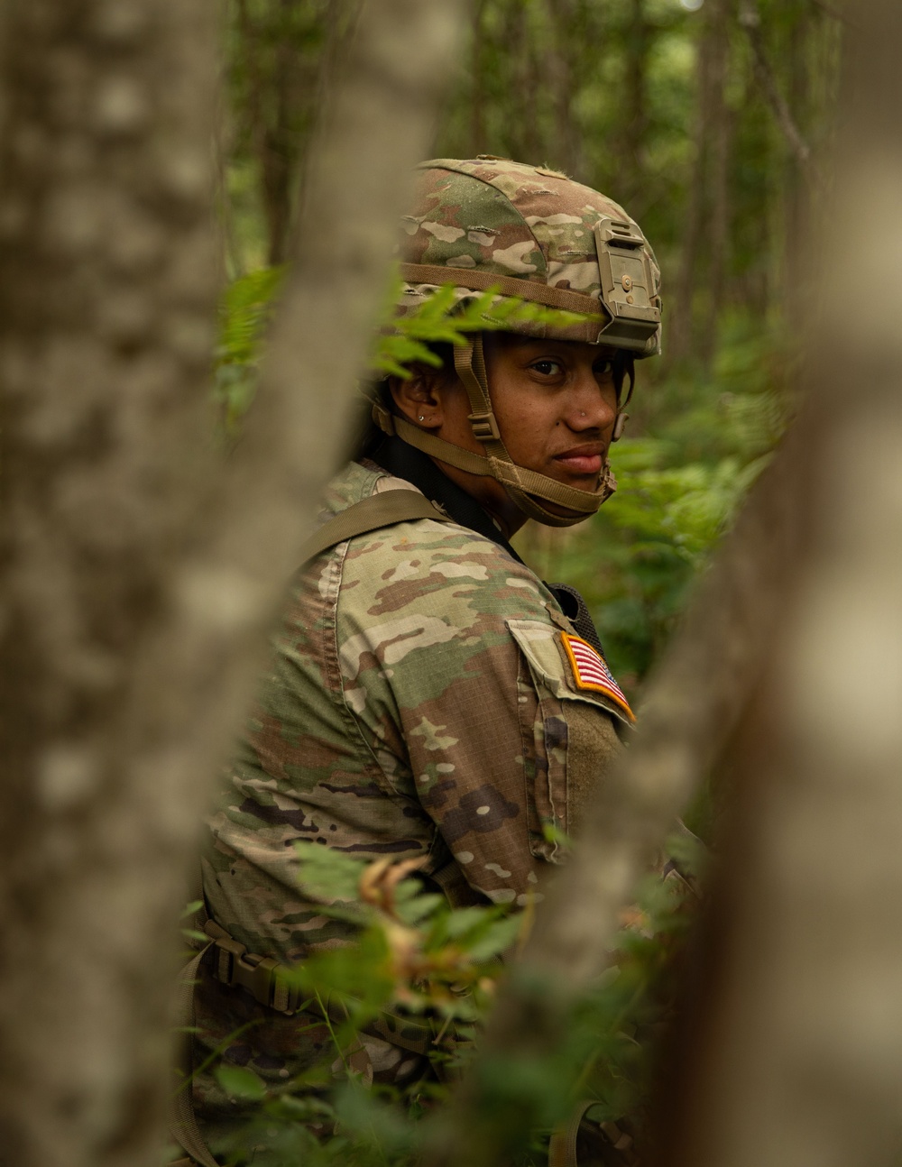 A U.S. Army soldier in full Operational Camouflage Pattern (OCP) uniform is photographed in a forested environment, looking back over their shoulder toward the camera. The soldier is equipped with a tactical helmet featuring a night-vision device mount and is wearing body armor with a reverse U.S. flag patch on the right sleeve. The image is captured through partially obscuring foreground foliage, creating a depth-of-field effect.