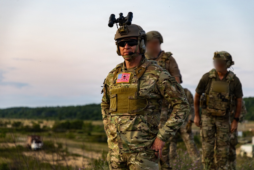 A U.S. Army soldier in full tactical gear stands in the foreground of an outdoor field environment, wearing OCP (Operational Camouflage Pattern) uniform with a visible American flag patch and 'ARMY' tape. Two additional military personnel with blurred/obscured faces are visible in the background. The setting appears to be a training exercise or operational deployment during dusk or dawn conditions.