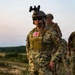 A U.S. Army soldier in full tactical gear stands in the foreground of an outdoor field environment, wearing OCP (Operational Camouflage Pattern) uniform with a visible American flag patch and 'ARMY' tape. Two additional military personnel with blurred/obscured faces are visible in the background. The setting appears to be a training exercise or operational deployment during dusk or dawn conditions.