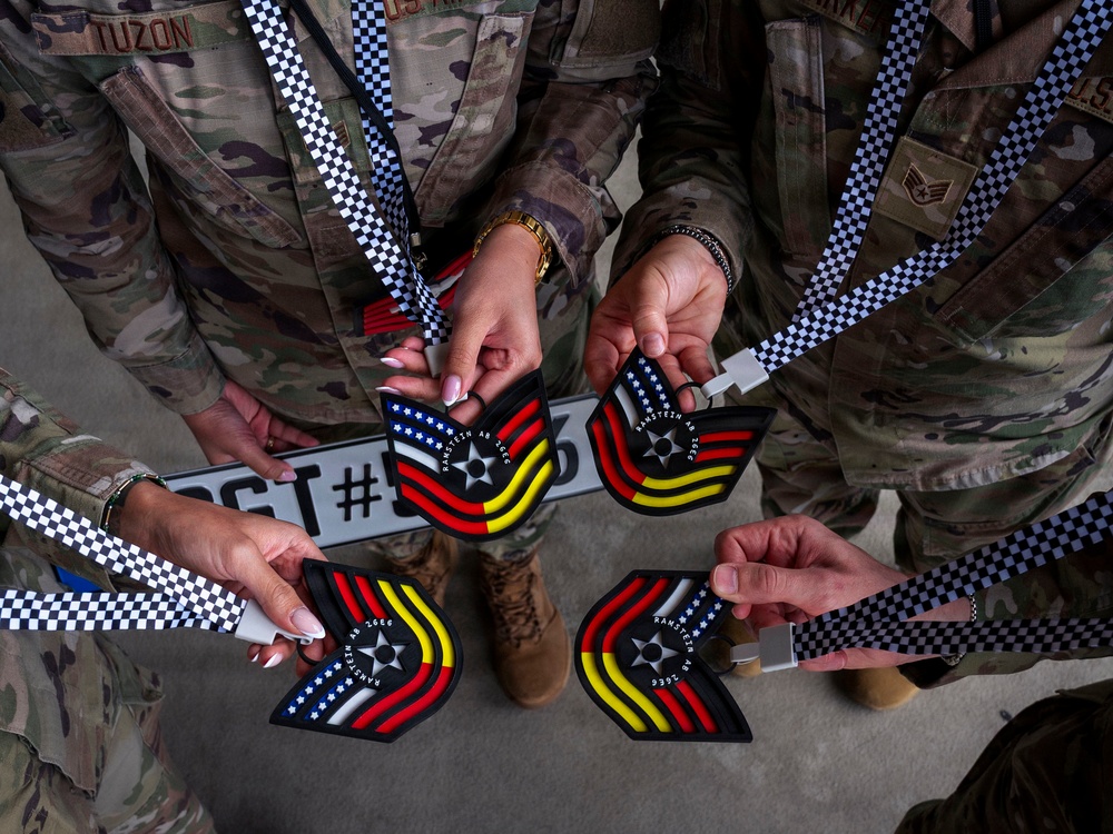 Military personnel in US Army/Air Force OCP (Operational Camouflage Pattern) uniforms are holding medals or commemorative medallions on black-and-white checkered lanyards. The medallions appear to be from Ramstein Air Base (AB 2666) and feature a chevron-shaped design incorporating the American flag stripes and German flag colors (black, red, yellow). A license plate-style placard with 'SGT #5__3' is also visible in the center of the frame.