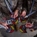 Military personnel in US Army/Air Force OCP (Operational Camouflage Pattern) uniforms are holding medals or commemorative medallions on black-and-white checkered lanyards. The medallions appear to be from Ramstein Air Base (AB 2666) and feature a chevron-shaped design incorporating the American flag stripes and German flag colors (black, red, yellow). A license plate-style placard with 'SGT #5__3' is also visible in the center of the frame.