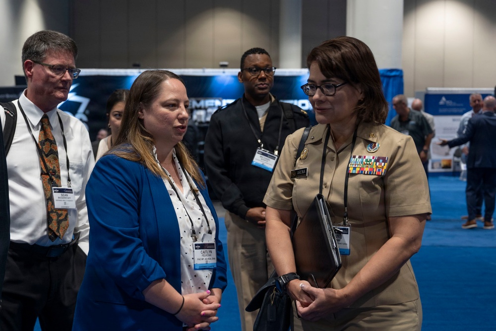 The image shows a professional conference or expo setting where several individuals are engaged in conversation. A woman in a U.S. military uniform with ribbons and insignia is speaking with a civilian woman wearing a blue blazer and a conference badge. Additional attendees and exhibition booths are visible in the background.