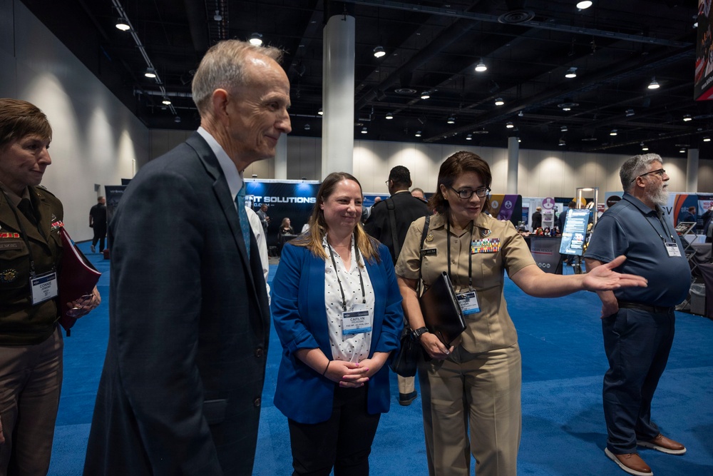 A group of individuals are engaged in conversation at what appears to be a military or defense industry conference/expo. The group includes at least two uniformed military personnel, civilian attendees wearing conference badges, and a man in a suit. The setting is a large convention hall with vendor booths visible in the background.