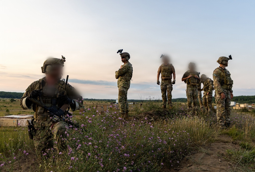 A group of military personnel in full combat gear are positioned on elevated terrain during dusk or dawn conditions. The individuals are wearing camouflage uniforms, tactical vests, and helmets equipped with night-vision device mounts. Their faces have been deliberately blurred, suggesting operational security considerations. A training range or mock facility is visible in the background.