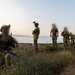 A group of military personnel in full combat gear are positioned on elevated terrain during dusk or dawn conditions. The individuals are wearing camouflage uniforms, tactical vests, and helmets equipped with night-vision device mounts. Their faces have been deliberately blurred, suggesting operational security considerations. A training range or mock facility is visible in the background.