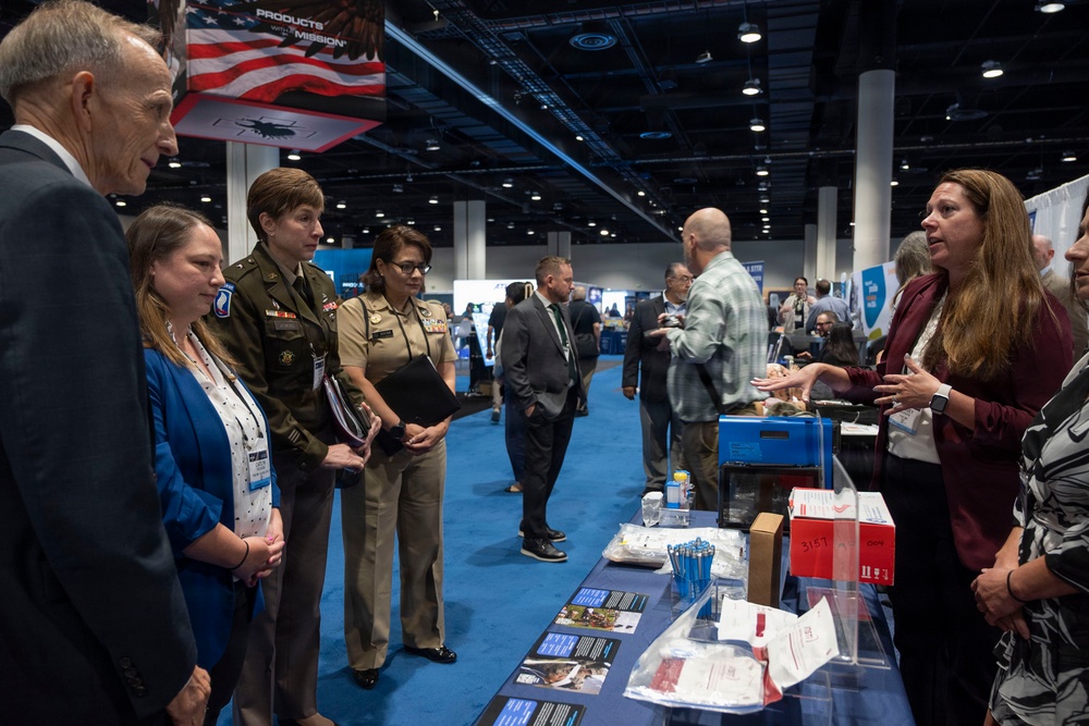 A military and civilian conference or trade show is taking place in a large convention center. Two uniformed military personnel — one in Army green service uniform and one in a tan/khaki naval or Marine uniform — are being briefed at an exhibit booth by a civilian woman in a burgundy blazer. The booth displays various materials, brochures, and small items on a blue-draped table.