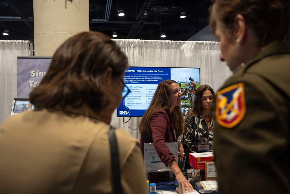 The image shows a Defense Health Agency (DHA) exhibition booth at what appears to be a military or defense-related conference or trade show. A presentation screen displays information about 'Warfighter Protection and Acute Care,' while booth staff interact with attendees including uniformed military personnel. The booth features informational materials, product displays, and signage related to military medical solutions.