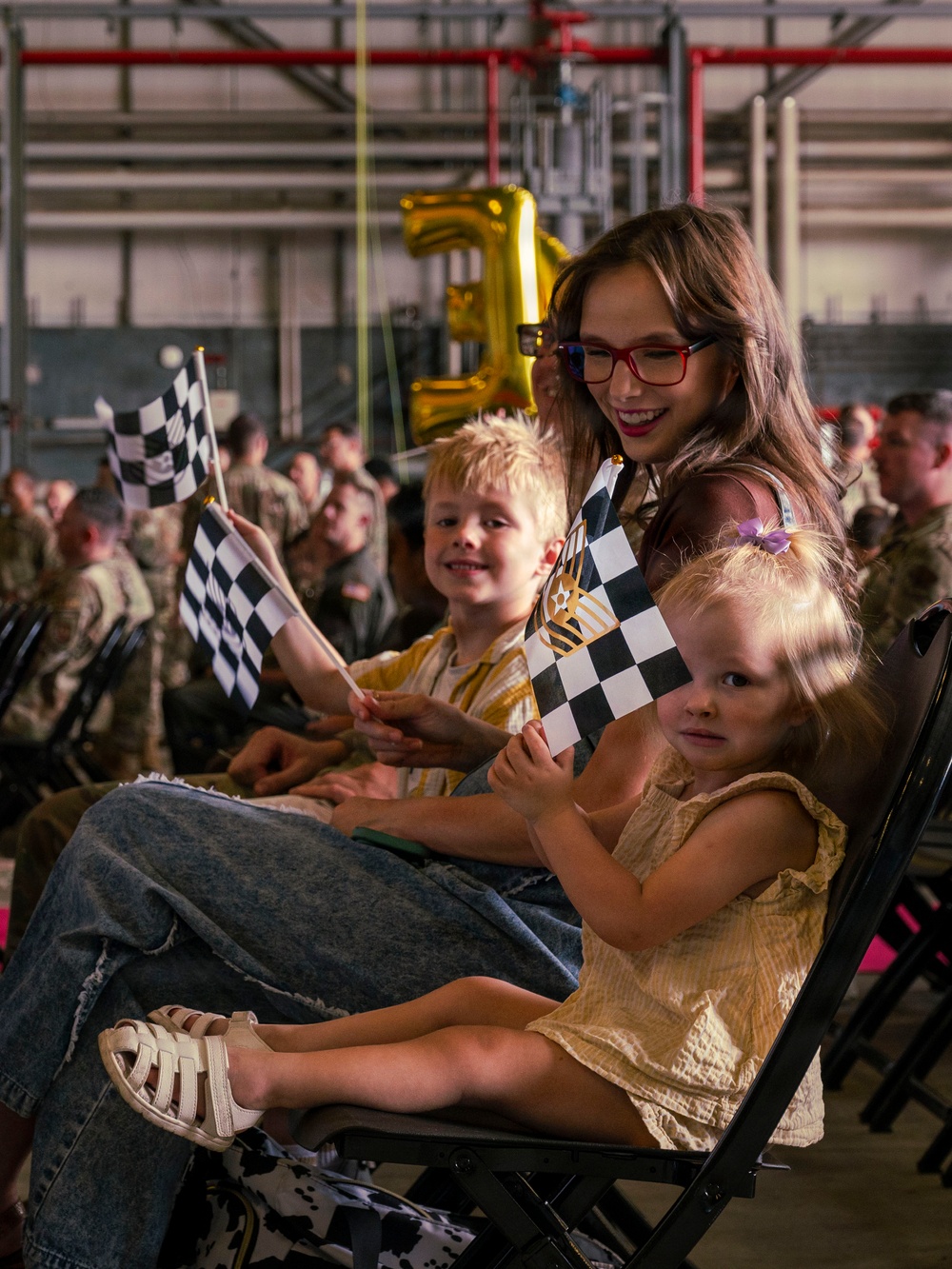 A young woman wearing red glasses sits with two small children at what appears to be a military ceremony or homecoming event held inside a hangar or large indoor facility. The group is holding black-and-white checkered flags, and gold number balloons are visible in the background. Rows of uniformed military personnel are seated in the background.