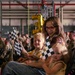 A young woman wearing red glasses sits with two small children at what appears to be a military ceremony or homecoming event held inside a hangar or large indoor facility. The group is holding black-and-white checkered flags, and gold number balloons are visible in the background. Rows of uniformed military personnel are seated in the background.