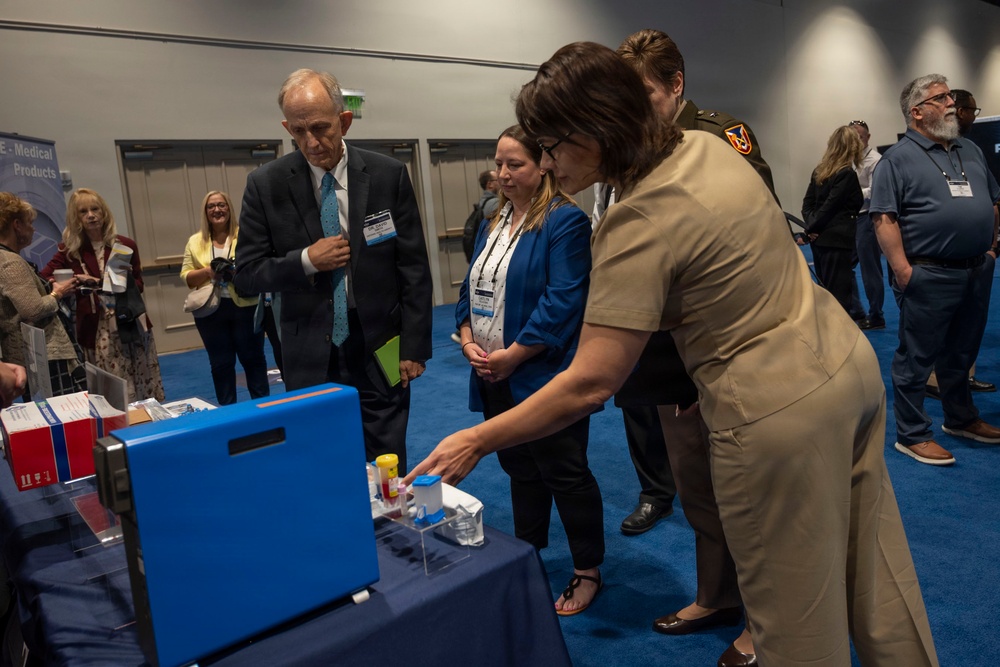 A conference or trade show setting where a person in military uniform is demonstrating medical products at a display table to a small group of attendees. The scene appears to be a professional medical or defense-related exhibition hall with multiple booths and attendees visible in the background. Participants are wearing name badge lanyards, indicating a formal conference environment.