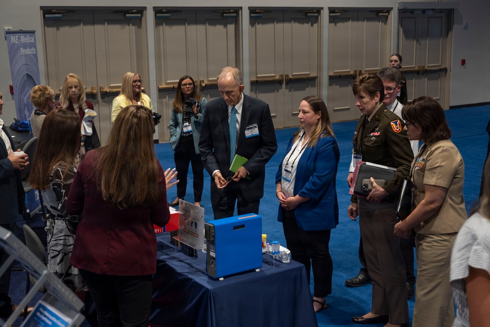 A professional conference or trade show event where attendees are gathered around an exhibit table displaying medical or technical equipment. The group includes civilians in business attire and at least two military personnel in uniform, suggesting a defense or military medical procurement event. A banner in the background reads 'PAE - Medical Products', indicating the exhibitor's identity.