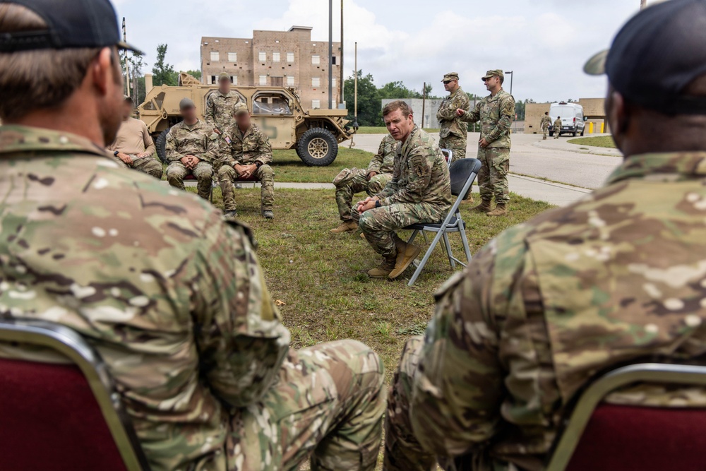 A group of military personnel in MultiCam/OCP camouflage uniforms are gathered outdoors on a grass area at what appears to be a military installation or training facility. One soldier is seated on a folding chair in a forward-leaning posture, appearing to lead or participate in a discussion or briefing with the group. A military tactical vehicle (HMMWV/Humvee variant) and multi-story training buildings are visible in the background.