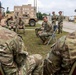 A group of military personnel in MultiCam/OCP camouflage uniforms are gathered outdoors on a grass area at what appears to be a military installation or training facility. One soldier is seated on a folding chair in a forward-leaning posture, appearing to lead or participate in a discussion or briefing with the group. A military tactical vehicle (HMMWV/Humvee variant) and multi-story training buildings are visible in the background.