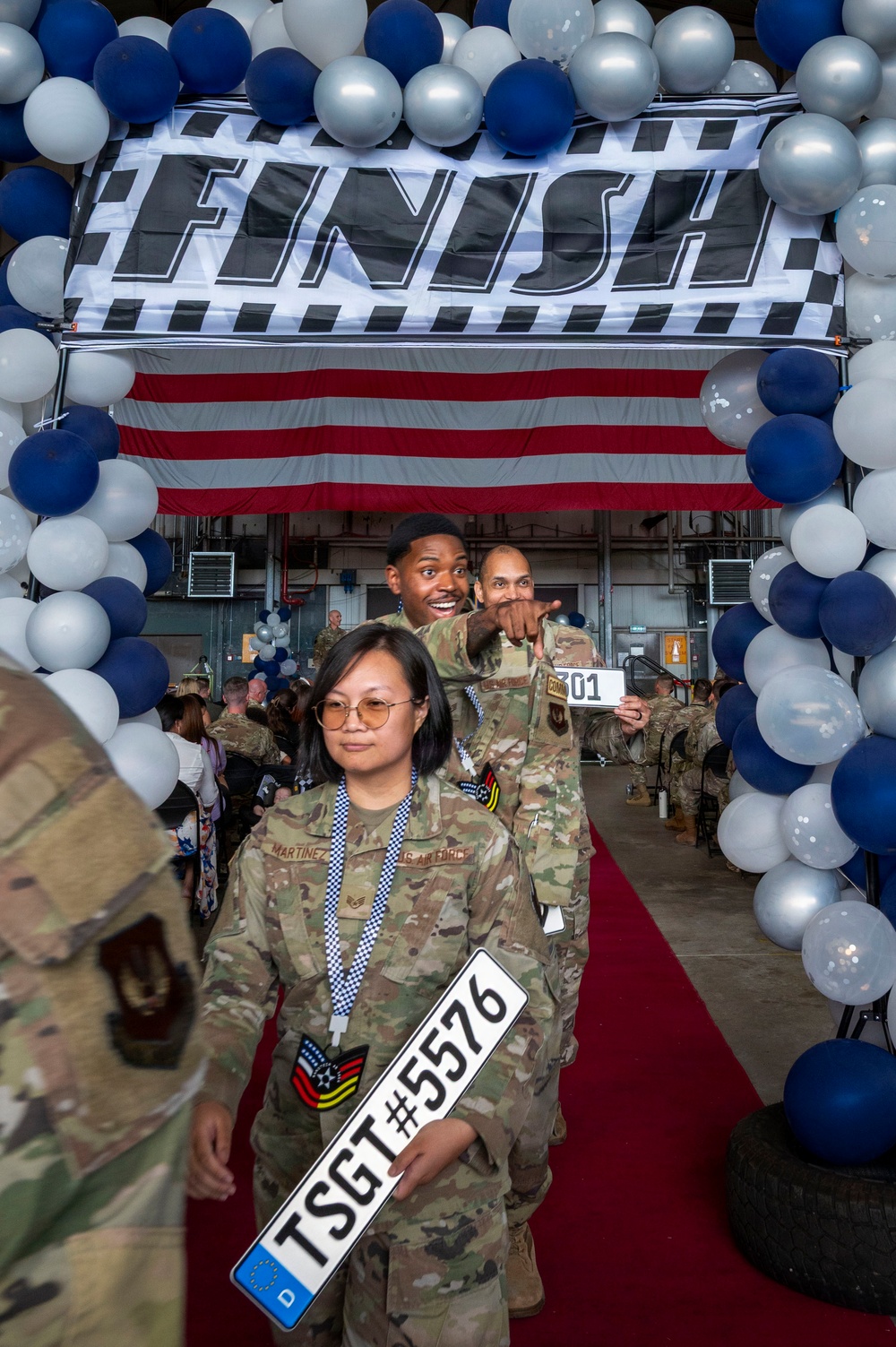 A military promotion ceremony with a racing theme, taking place inside what appears to be an aircraft hangar. Personnel in US Air Force OCP (Operational Camouflage Pattern) uniforms walk under a balloon arch and 'FINISH' banner on a red carpet, holding license plate-style signs indicating their new rank. The foreground airman holds a sign reading 'TSGT#5576', indicating promotion to Technical Sergeant.