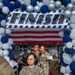 A military promotion ceremony with a racing theme, taking place inside what appears to be an aircraft hangar. Personnel in US Air Force OCP (Operational Camouflage Pattern) uniforms walk under a balloon arch and 'FINISH' banner on a red carpet, holding license plate-style signs indicating their new rank. The foreground airman holds a sign reading 'TSGT#5576', indicating promotion to Technical Sergeant.