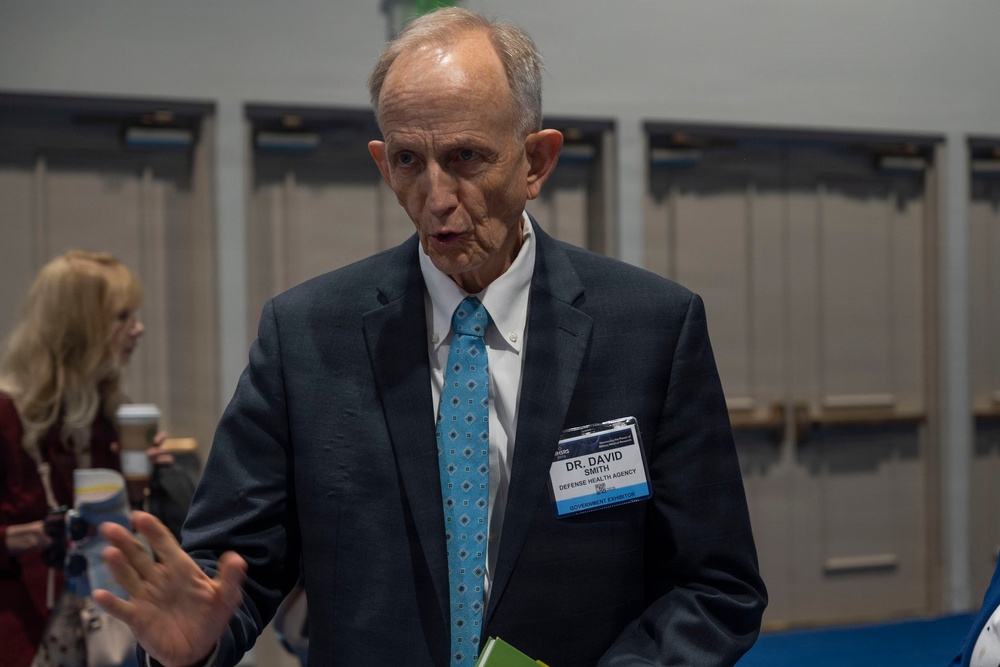 A man identified by his name badge as Dr. David Smith from the Defense Health Agency is speaking at what appears to be a professional conference or expo. He is wearing a dark suit with a light blue patterned tie and gesturing with his hand while talking. A woman with blonde hair is visible in the background, also at the event.