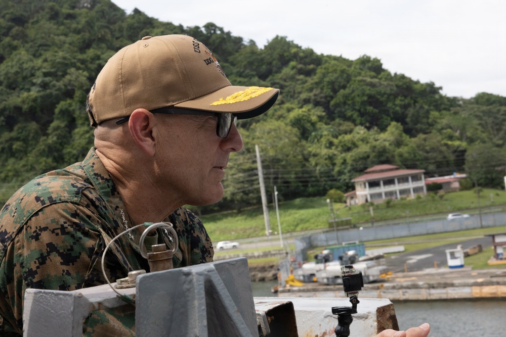 A senior military officer wearing USMC digital camouflage (MARPAT) uniform and a tan cap with gold scrambled eggs insignia is observing from what appears to be a vessel or dock structure. The setting appears to be a tropical waterway or port, with lush green hills, a colonial-style building, and watercraft visible in the background. A small action camera is mounted nearby on a rail.