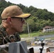 A senior military officer wearing USMC digital camouflage (MARPAT) uniform and a tan cap with gold scrambled eggs insignia is observing from what appears to be a vessel or dock structure. The setting appears to be a tropical waterway or port, with lush green hills, a colonial-style building, and watercraft visible in the background. A small action camera is mounted nearby on a rail.