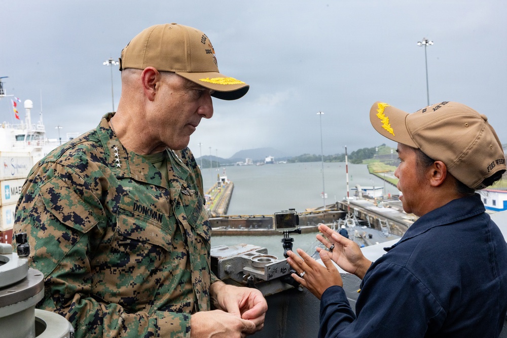 Two military personnel are engaged in conversation aboard what appears to be a naval vessel transiting through a canal lock, likely the Panama Canal. The individual on the left wears U.S. Marine Corps MARPAT camouflage uniform with the name tape 'DONOVAN' and three-star rank insignia indicating a Lieutenant General. Both individuals wear matching tan/coyote brown baseball caps with yellow insignia.