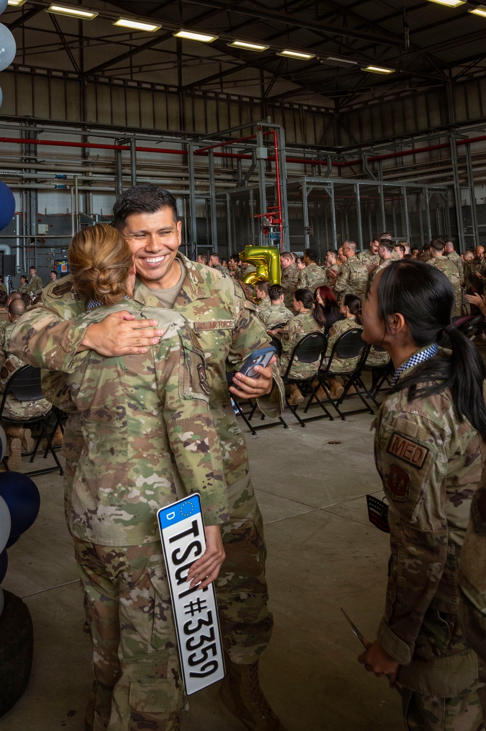 A U.S. Air Force military ceremony taking place inside a large hangar facility. A smiling male Airman in OCP (Operational Camouflage Pattern) uniform is embracing a female service member while holding a German vehicle registration plate reading 'TSU #3359'. A large group of uniformed personnel are seated in the background near gold balloon numbers.