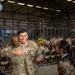 A U.S. Air Force military ceremony taking place inside a large hangar facility. A smiling male Airman in OCP (Operational Camouflage Pattern) uniform is embracing a female service member while holding a German vehicle registration plate reading 'TSU #3359'. A large group of uniformed personnel are seated in the background near gold balloon numbers.
