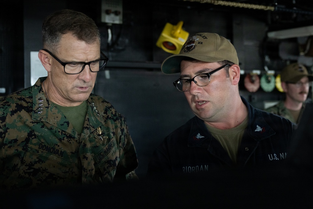 Two military personnel are engaged in a discussion, appearing to review something together. The individual on the left wears U.S. Marines digital camouflage with the name tape 'DONOVAN' and visible rank insignia suggesting a senior officer. The individual on the right wears a U.S. Navy uniform with the name tape 'RIORDAN' and a tan cap with what appears to be a ship designation.