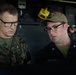 Two military personnel are engaged in a discussion, appearing to review something together. The individual on the left wears U.S. Marines digital camouflage with the name tape 'DONOVAN' and visible rank insignia suggesting a senior officer. The individual on the right wears a U.S. Navy uniform with the name tape 'RIORDAN' and a tan cap with what appears to be a ship designation.