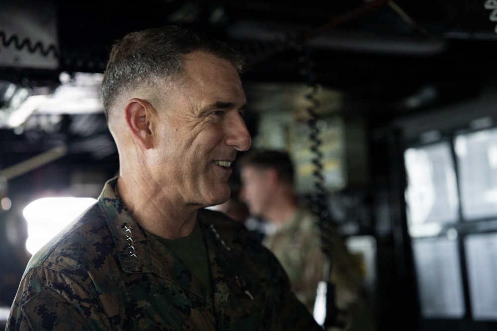 A senior U.S. Marine Corps officer wearing MARPAT camouflage uniform is visible in profile, smiling, aboard what appears to be a naval vessel or military facility. The officer displays four stars on the collar, indicating the rank of General. A second military personnel member is visible in the blurred background.
