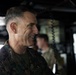 A senior U.S. Marine Corps officer wearing MARPAT camouflage uniform is visible in profile, smiling, aboard what appears to be a naval vessel or military facility. The officer displays four stars on the collar, indicating the rank of General. A second military personnel member is visible in the blurred background.