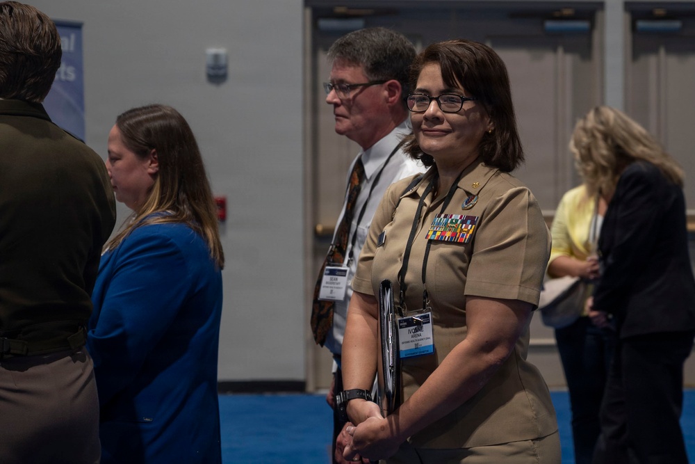 The image shows a group of individuals at what appears to be a professional conference or military event. A woman in U.S. Navy tan service uniform with multiple ribbons and a conference badge is prominently featured in the foreground. Several other attendees in civilian attire are visible in the background.