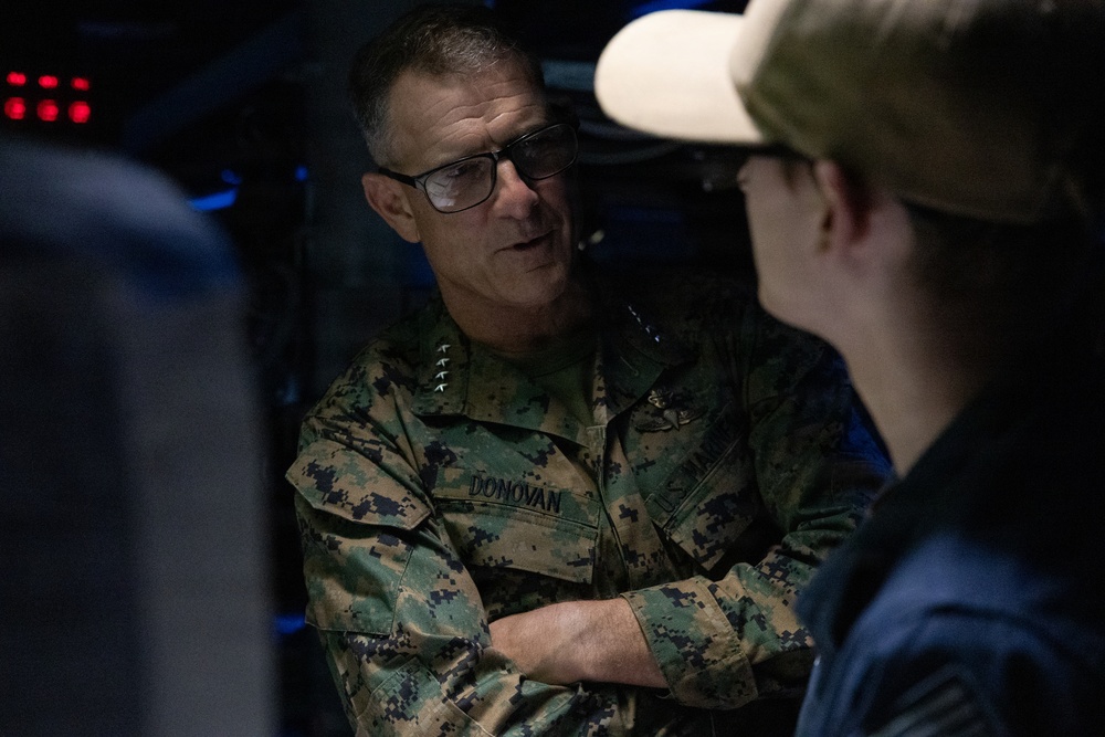 A U.S. Marine Corps officer with the name tape 'DONOVAN' is engaged in conversation in a dimly lit environment. He is wearing MARPAT camouflage uniform with general officer rank insignia (four stars visible on collar). He appears to be speaking with another individual whose back is partially visible in the foreground.